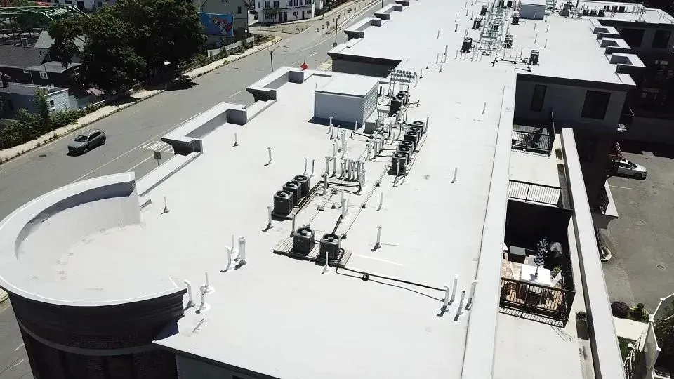 Aerial view of a flat commercial roof with HVAC units, antennas, and a curved building section. Gray building next to a street.