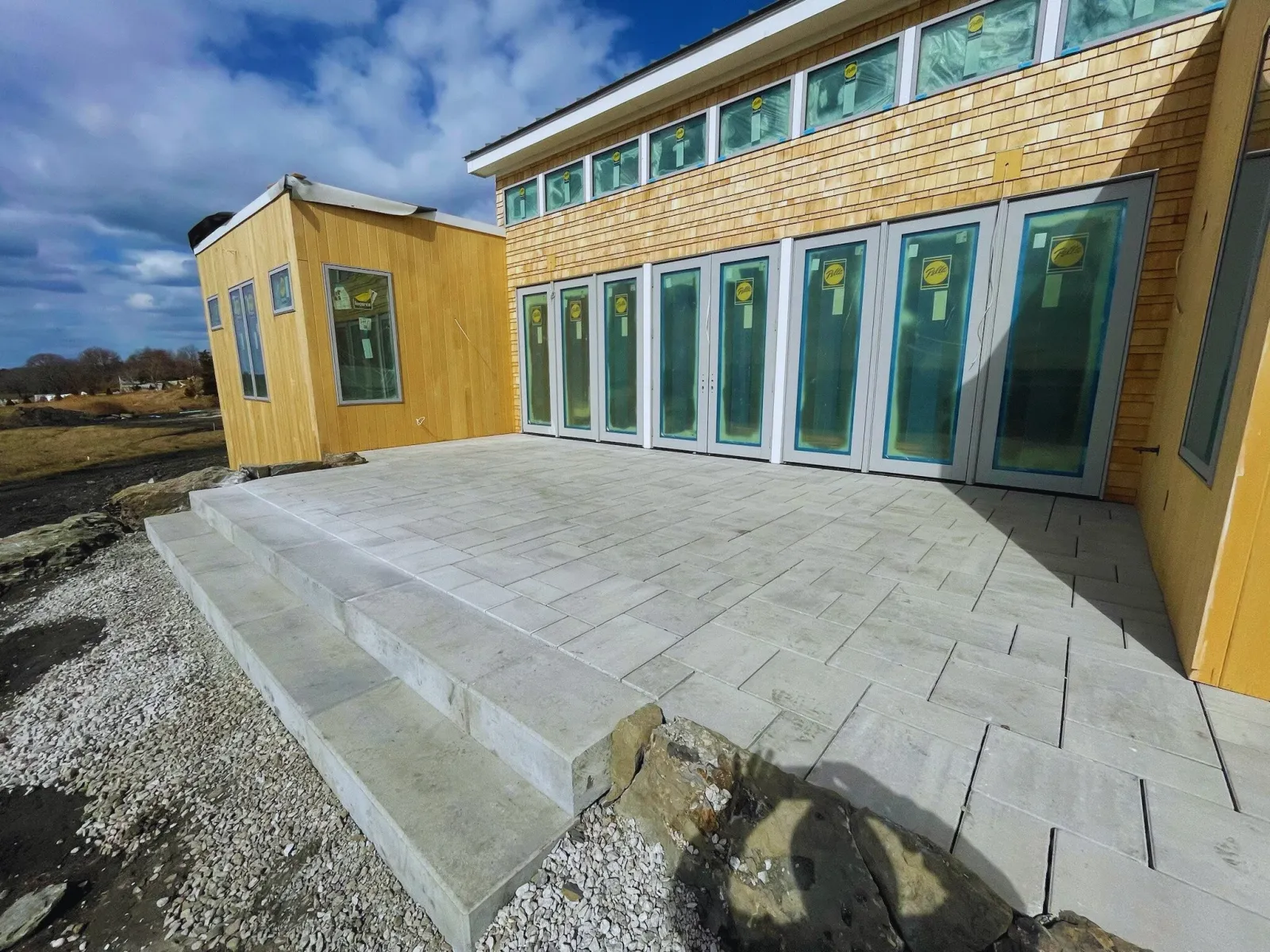 Modern building exterior with large windows, wooden siding, and a stone patio under a cloudy sky.