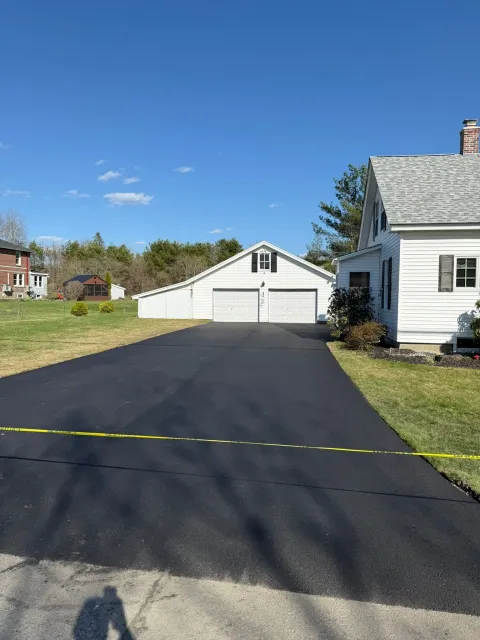 A freshly paved asphalt driveway leads to a two-car garage beside a white house on a sunny day.