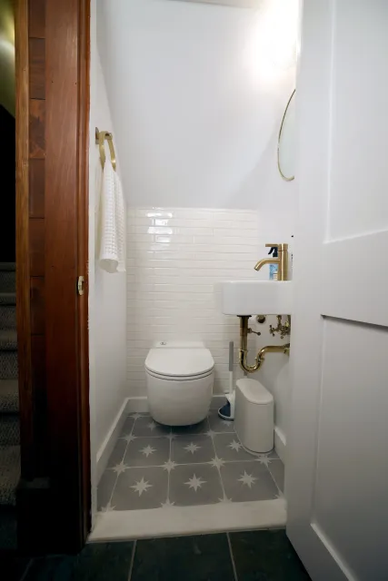 Small bathroom with a white toilet, a small wall-mounted sink with a gold faucet, a round mirror, a white waste bin, and a white wall with a beige towel hanging on a gold hook.