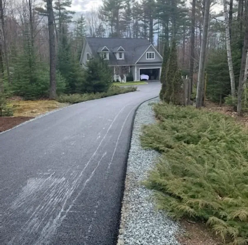 A long, paved driveway leads through a wooded area to a gray house with a garage and two dormer windows.