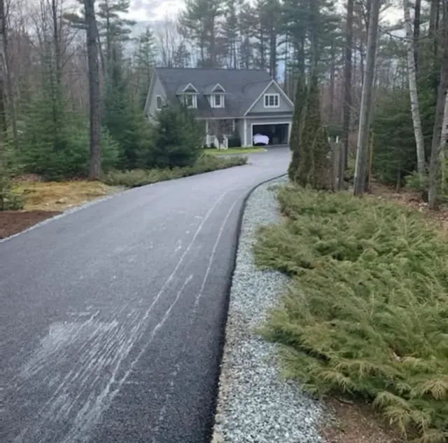 A long, paved driveway leads through a wooded area to a gray house with a garage and two dormer windows.