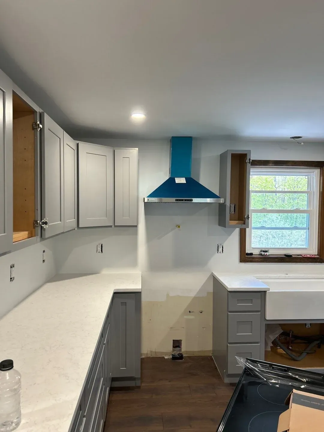 Kitchen under renovation with white cabinets, blue range hood, and a window above a small work area.