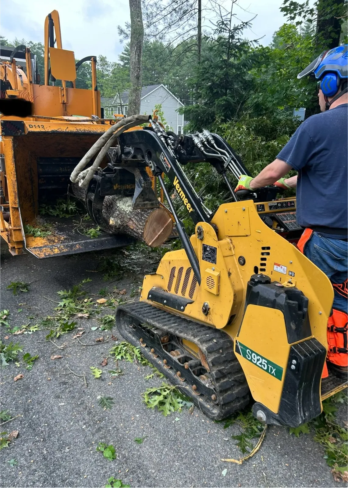 A worker operating a small yellow woodchipper with tracks, cutting fallen branches and leaves in a yard. The woodchipper is feeding a large cut tree log, while green tree branches and leaves are scattered around the ground.