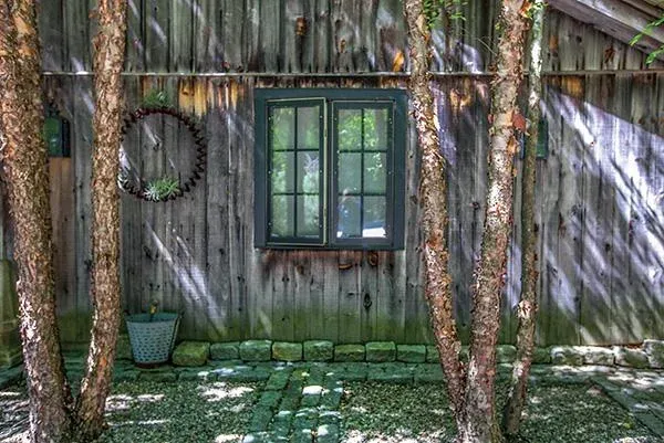 A weathered wooden fence with a small window, surrounded by tall trees with bark. A brick border lines the ground beneath, which is covered in grass, and a decorative bicycle wheel hangs on the fence.