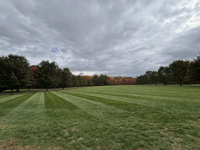 A large open grassy field with freshly mowed stripes, lined by rows of trees with green and autumn-colored leaves, under a cloudy sky.