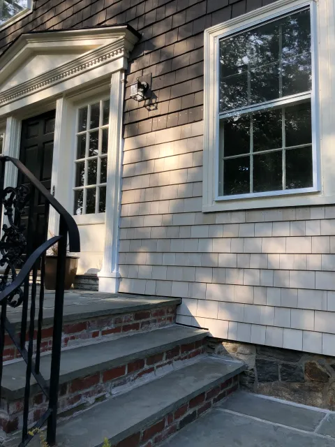 Front steps with brick and stone leading up to a house entrance with black door, white trim, a wall-mounted lantern, and a window reflecting trees.