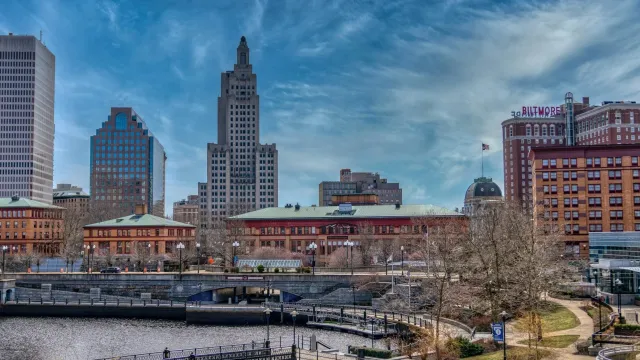 City skyline with tall buildings, a river, trees, and a park in front, under a partly cloudy sky.