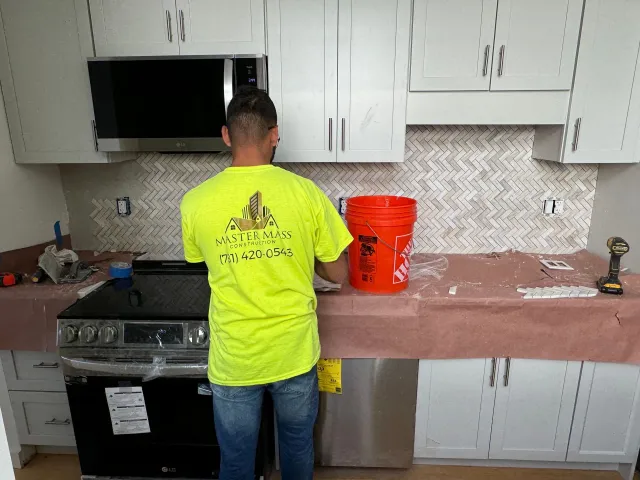 Man tiling a kitchen backsplash, standing in front of cabinets and a stove, holding a trowel next to a bucket.