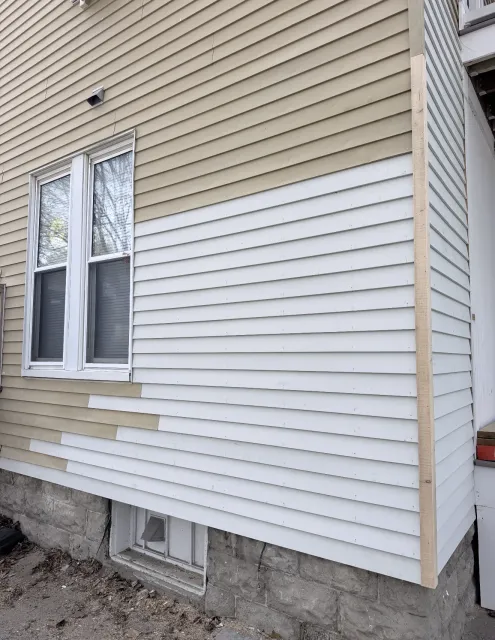 Side of a beige house with white vinyl siding, windows, and a small concrete foundation.