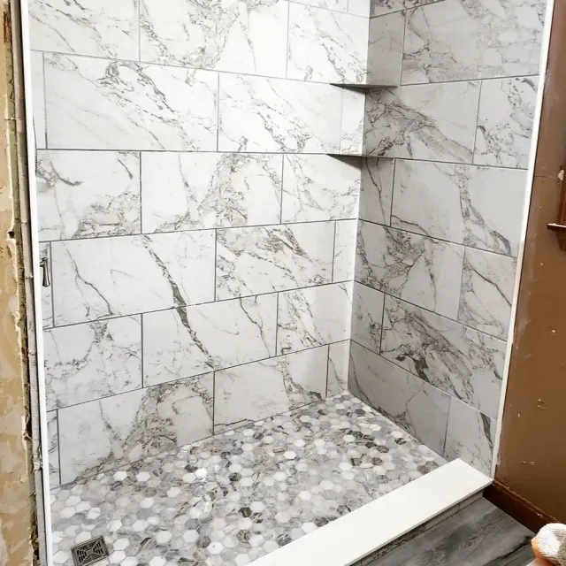 A finished walk-in shower with marble wall tiles and a pebble stone floor, featuring built-in corner shelves.