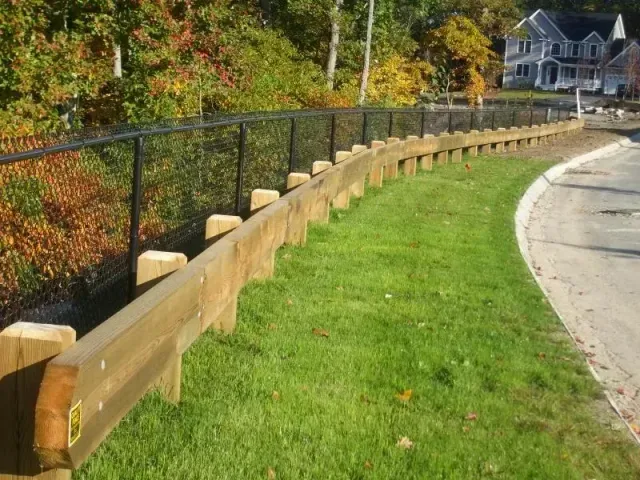 A wooden guardrail runs along a curved road, with a black chain-link fence behind it and trees in the background.