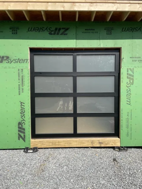 Black-framed glass garage door installed in a new building with green sheathing.