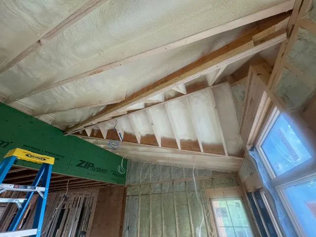 Partially insulated attic interior with exposed rafters, a blue ladder, and windows letting in daylight