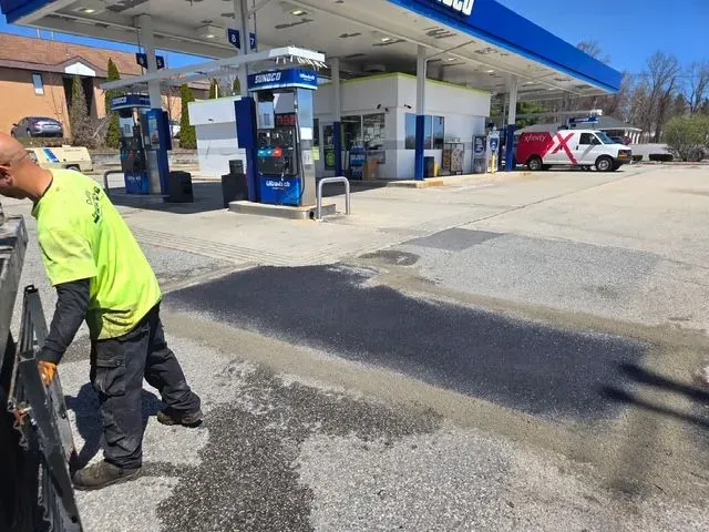 Gas station with a worker patching a dark asphalt repair near the fuel pumps.