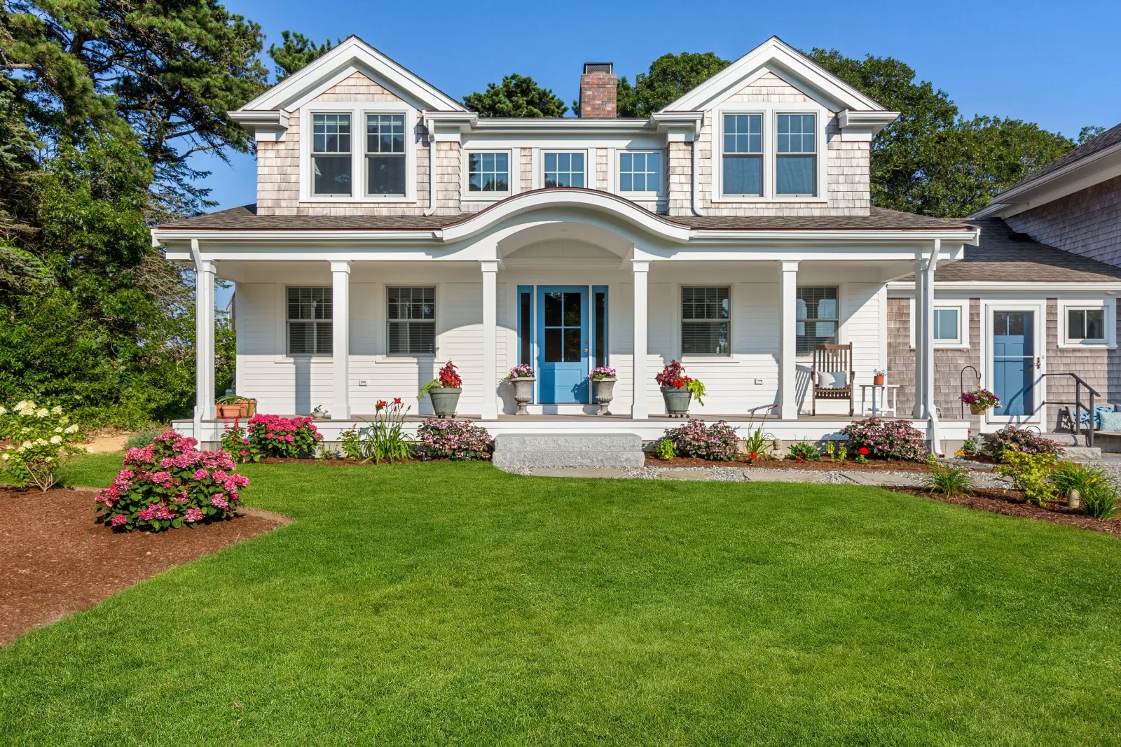 Front view of a two-story white house with a covered porch, blue front door, multiple windows, and a garden with colorful flowers in the foreground. Tall trees are visible behind the house under a clear blue sky.