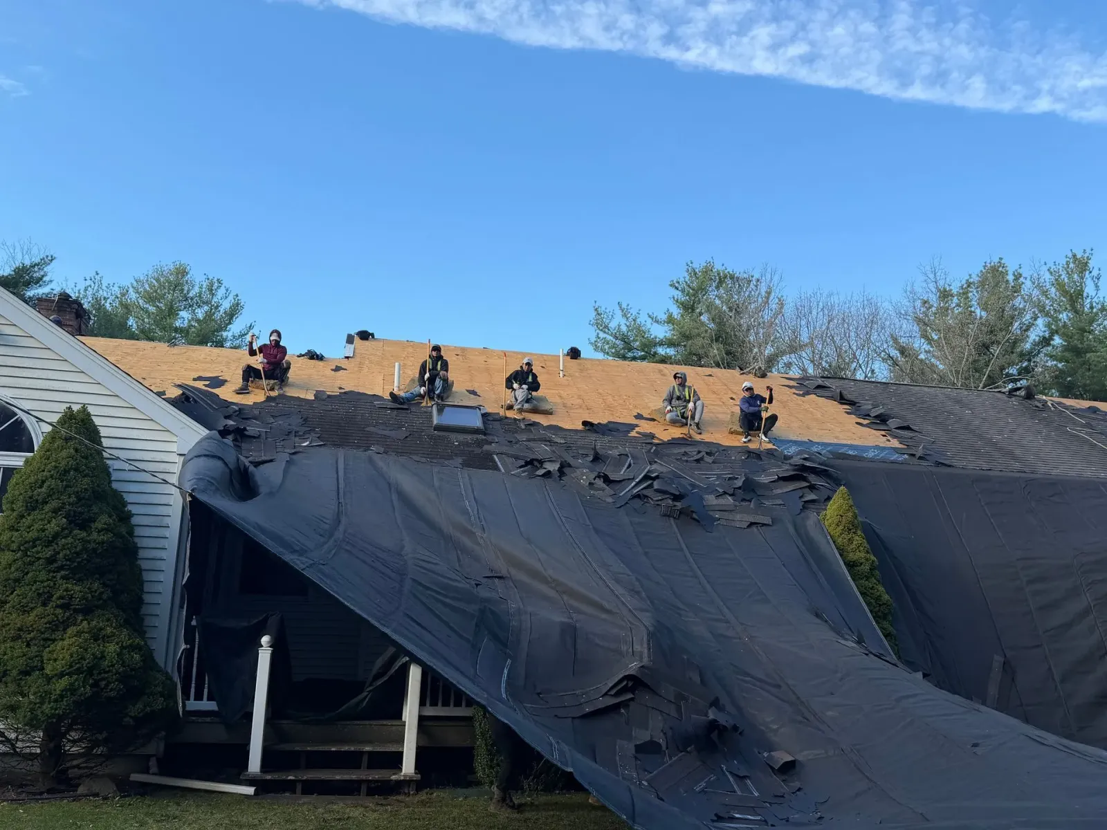 Roofers working on a house roof with black tarp and plywood under a blue sky