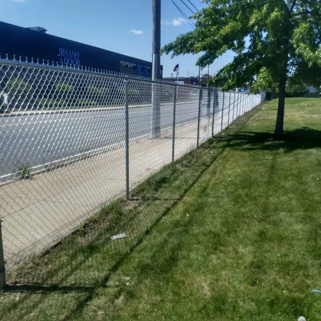 Chain link fence alongside a sidewalk and grassy area, with a building in the background.