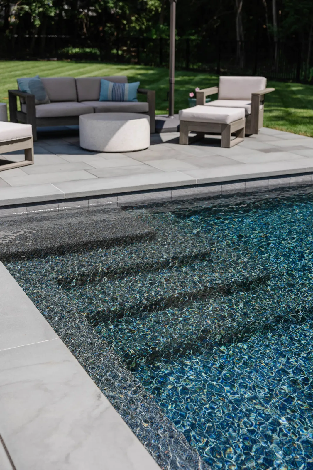 Outdoor pool with steps leading into the water, surrounded by grey stone tiles, with outdoor furniture including a sofa, chairs, and a round table on a patio with grass and trees in the background.