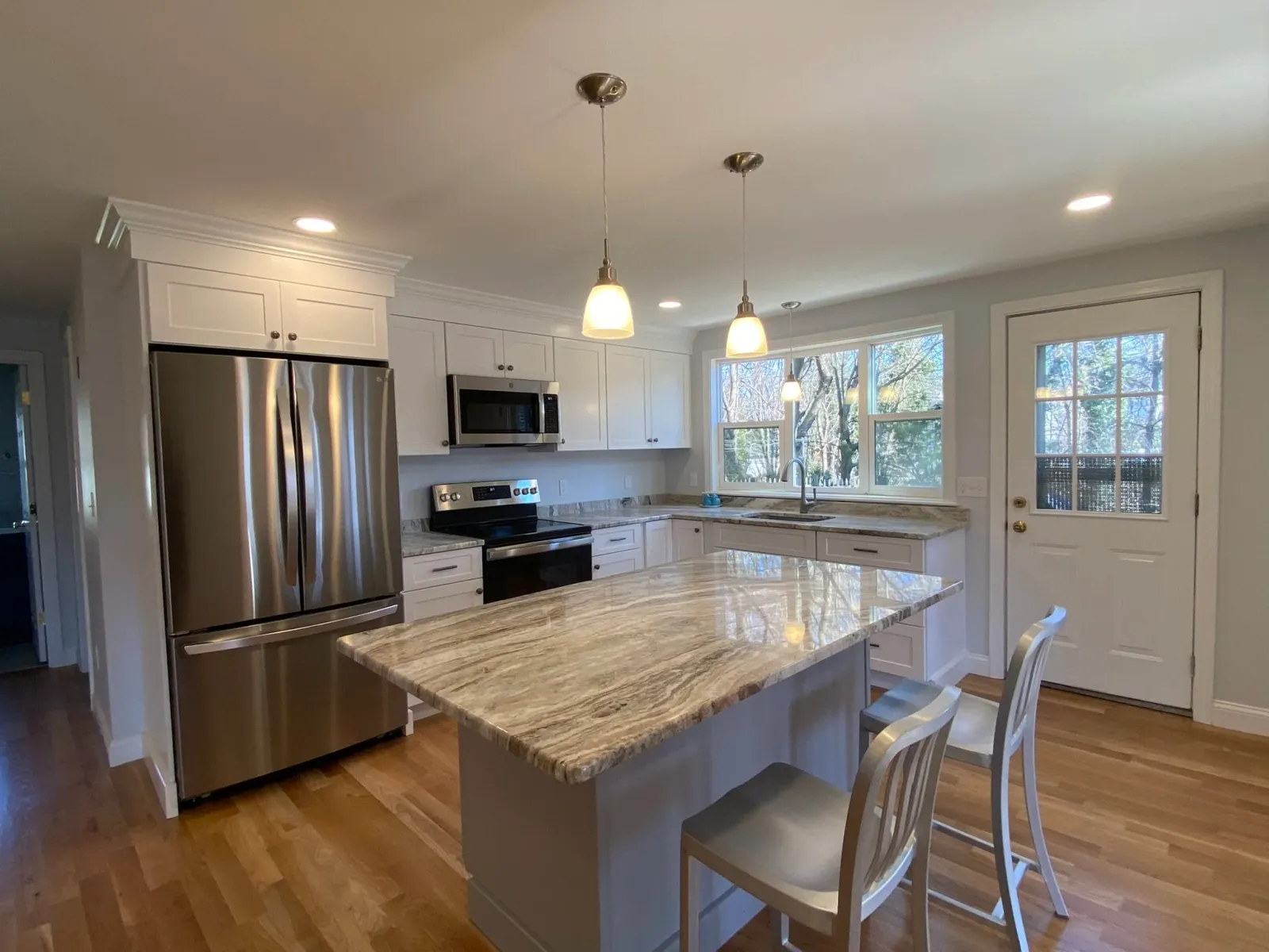 Modern kitchen with white cabinetry, stainless steel refrigerator, stove, microwave, granite countertops, island with seating, hardwood floors, large windows, and a door leading outside.