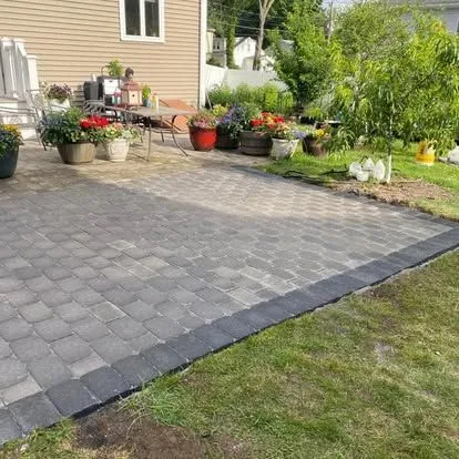 Paved patio beside a house with potted flowers and garden beds in a sunny yard.