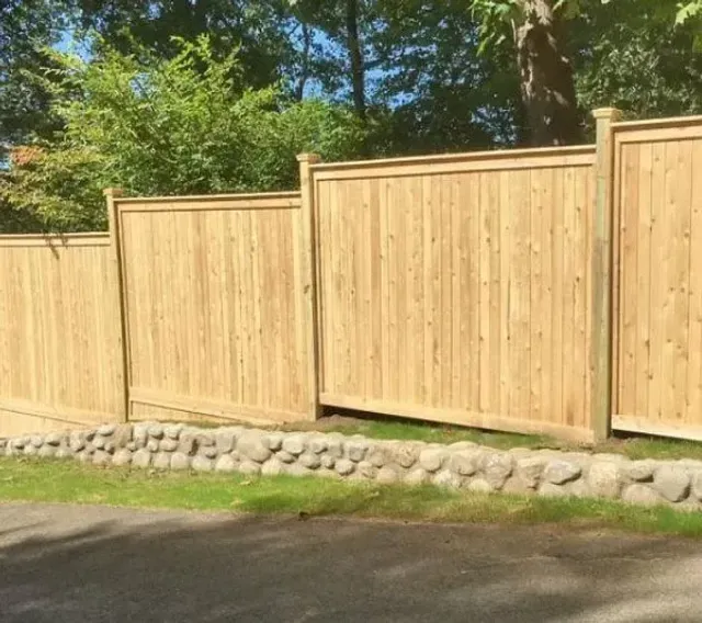 Wooden privacy fence along a road, with stone edging and trees behind it.