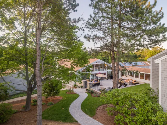 A backyard with a winding stone pathway, green shrubs, trees, and a patio area with outdoor furniture and a couple of people sitting at a table, in front of a house with large windows and a screened porch.