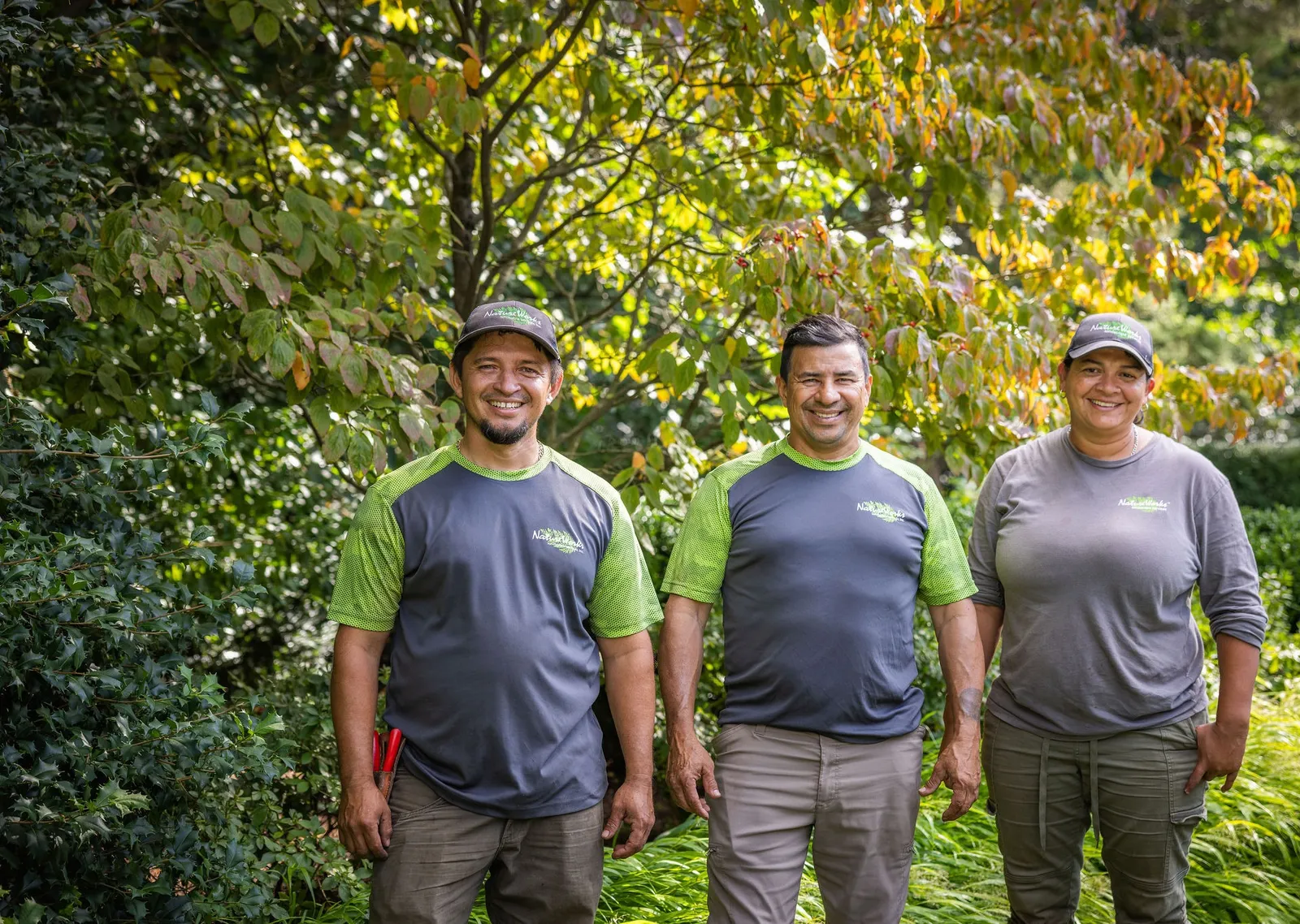 Three people stand smiling outdoors, wearing work uniforms with green accents, surrounded by lush greenery and plants in a garden setting.
