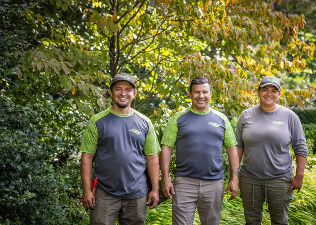 Three people stand smiling outdoors, wearing work uniforms with green accents, surrounded by lush greenery and plants in a garden setting.