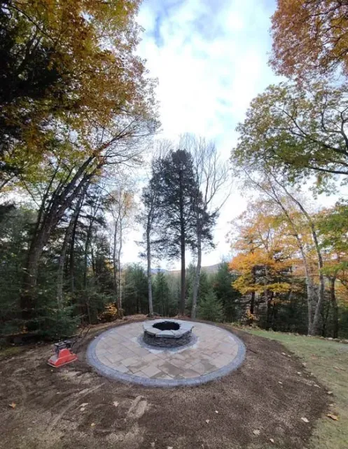 A circular stone fire pit sits in the center of a newly installed stone patio, surrounded by trees with autumn foliage.