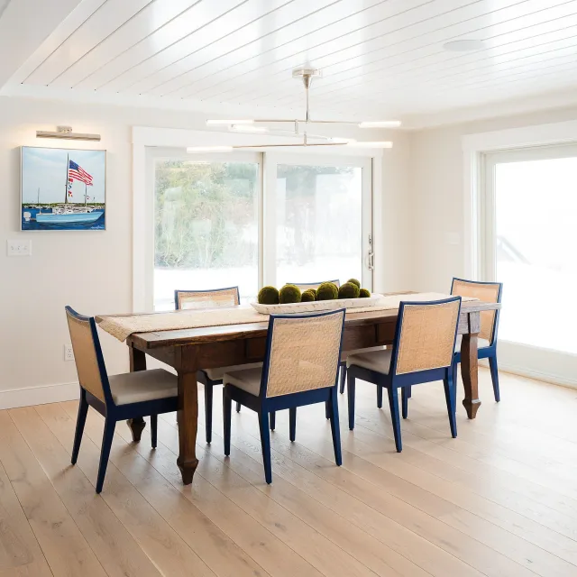 Hardwood floor installation in a Scituate dining room with a wooden table, six chairs, a modern chandelier, large windows, and nautical artwork featuring a boat with American flags.