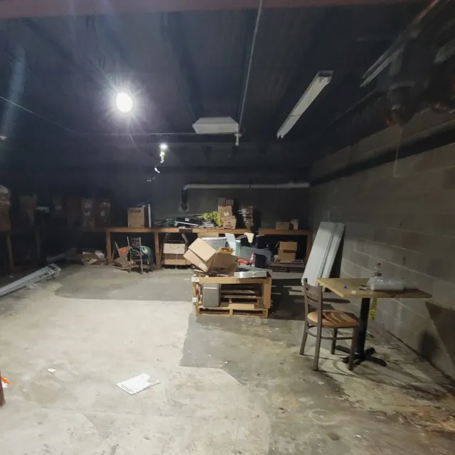 A cluttered storage room with boxes and miscellaneous items on shelves, a small table with a chair, and a plain concrete floor and brick wall.