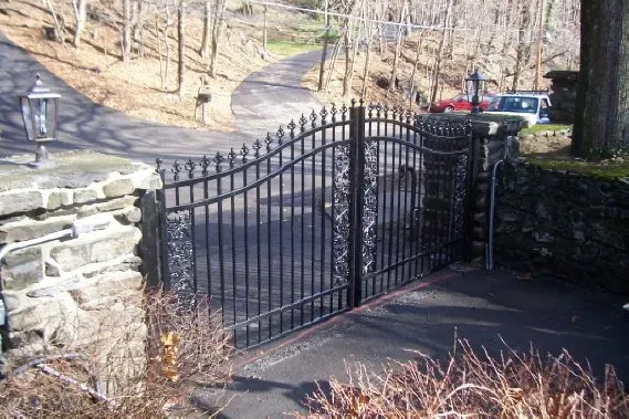 Black metal gate at the end of a stone wall on a paved driveway, with hillside and parked cars beyond