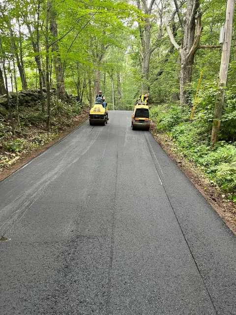 Two road rollers compacting fresh asphalt on a tree-lined road.