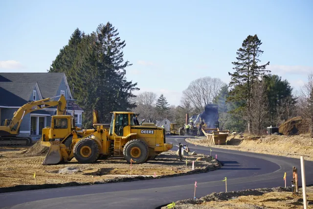 Construction site with yellow heavy machinery and workers laying asphalt on a curved road, with houses and tall pine trees in the background under a clear blue sky.