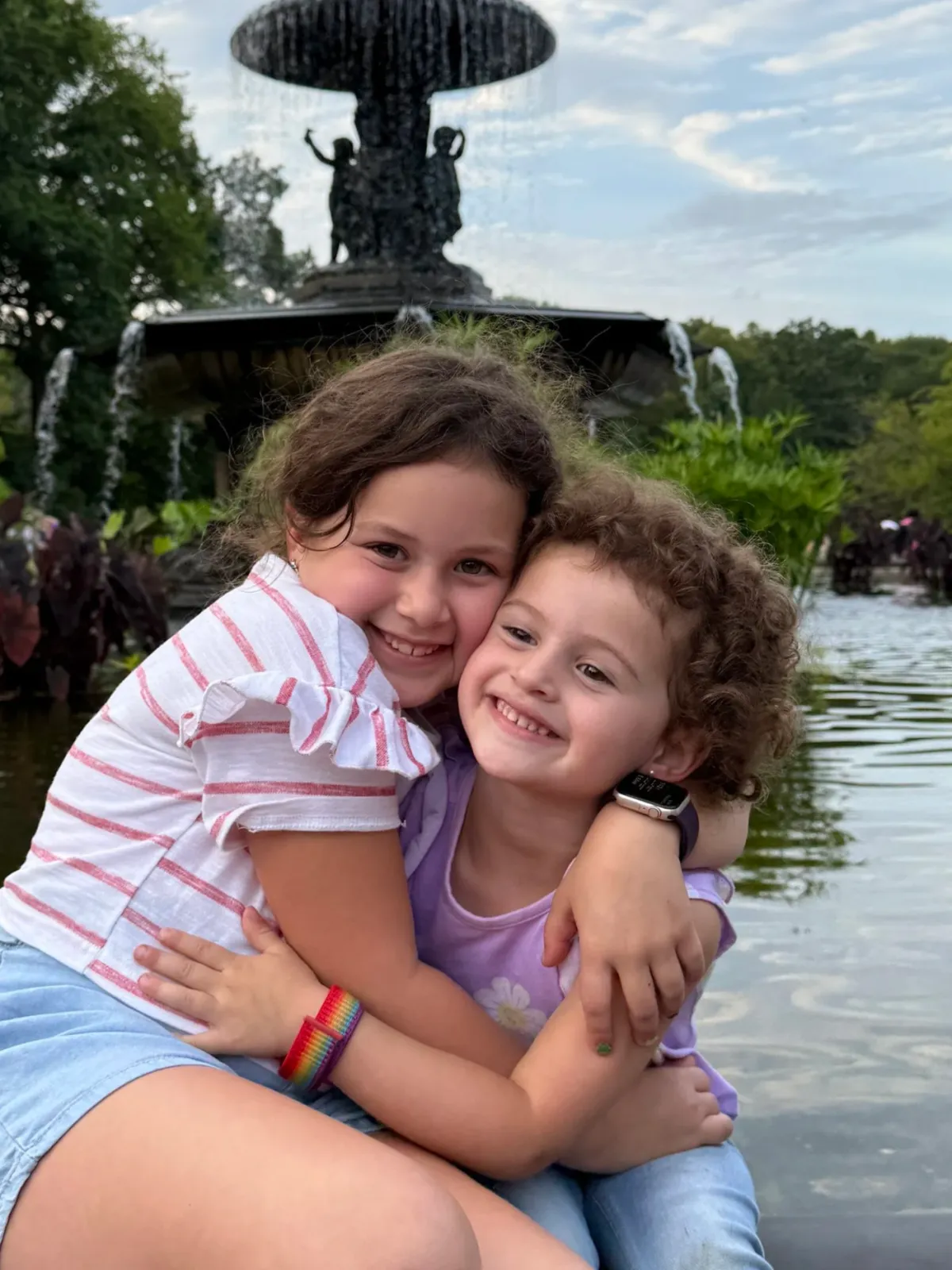 A split image showing two children smiling. The child on the left has curly light brown hair, freckles, and is holding a carousel ride at night. The child on the right has dark, shoulder-length curly hair and is smiling outdoors with a beach and ocean in the background.