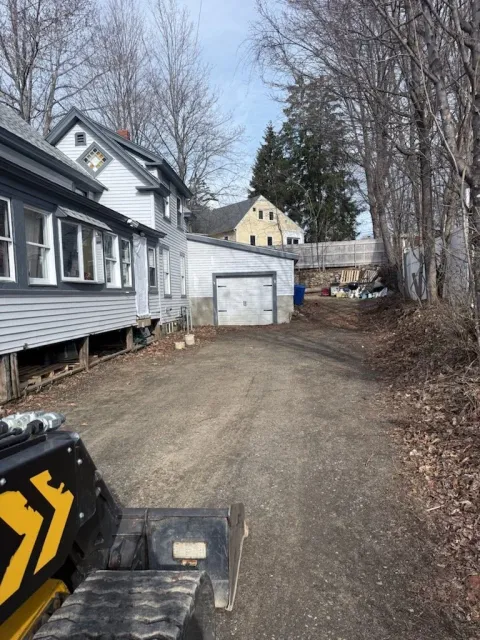 A gravel driveway in front of a white house with several windows and a garage, with leafless trees and a neighboring house in the background.