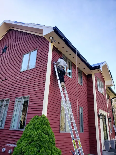 Professional contractor on a ladder performing exterior home repairs on a red farmhouse with vinyl siding.