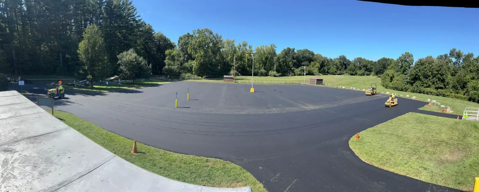A wide, freshly paved asphalt parking lot surrounded by grassy fields and trees under a clear blue sky.