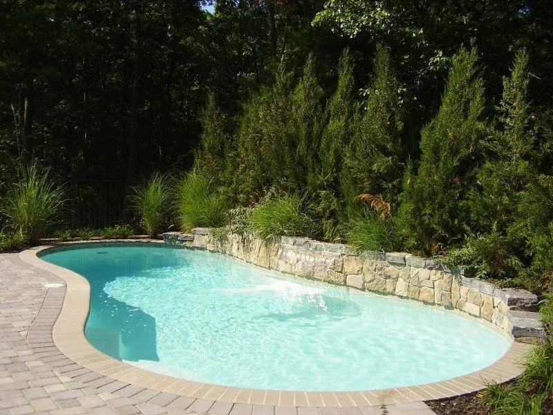 Above-ground swimming pool surrounded by brick paving and greenery, with a stone retaining wall and dense trees in the background.