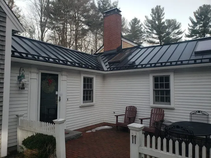 White house exterior with black metal roof, brick chimney, and small picket-fenced courtyard.
