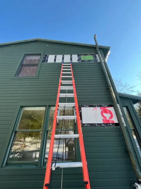 A two-story house with green siding and multiple large windows, with a tall red ladder leaning against it. There is construction or renovation work happening, as indicated by insulation and flashing tape around some windows, and a person taking a photography reflection in the lower window.