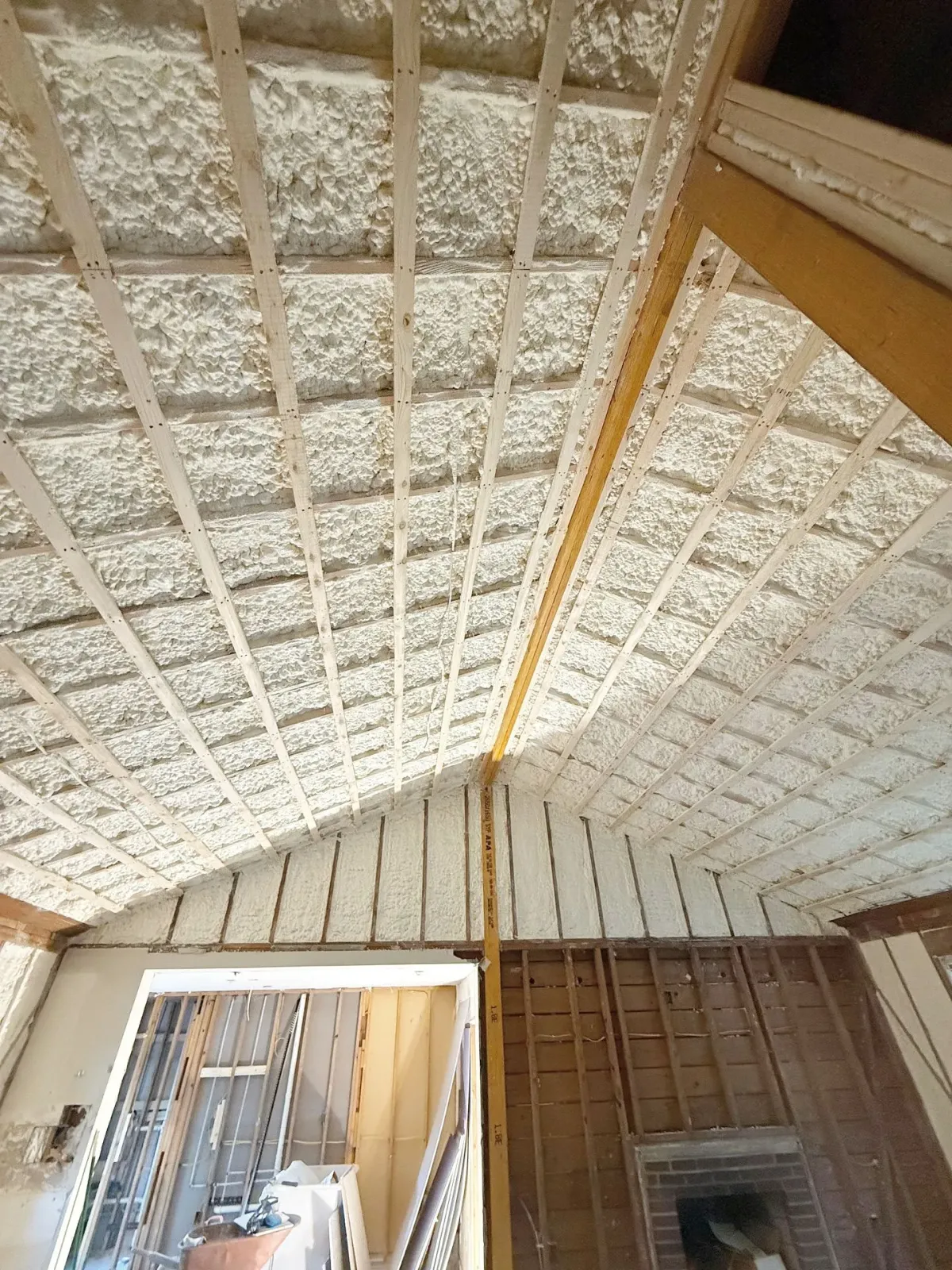 Interior view of a partially built room with spray foam insulation in the ceiling between exposed wood beams.
