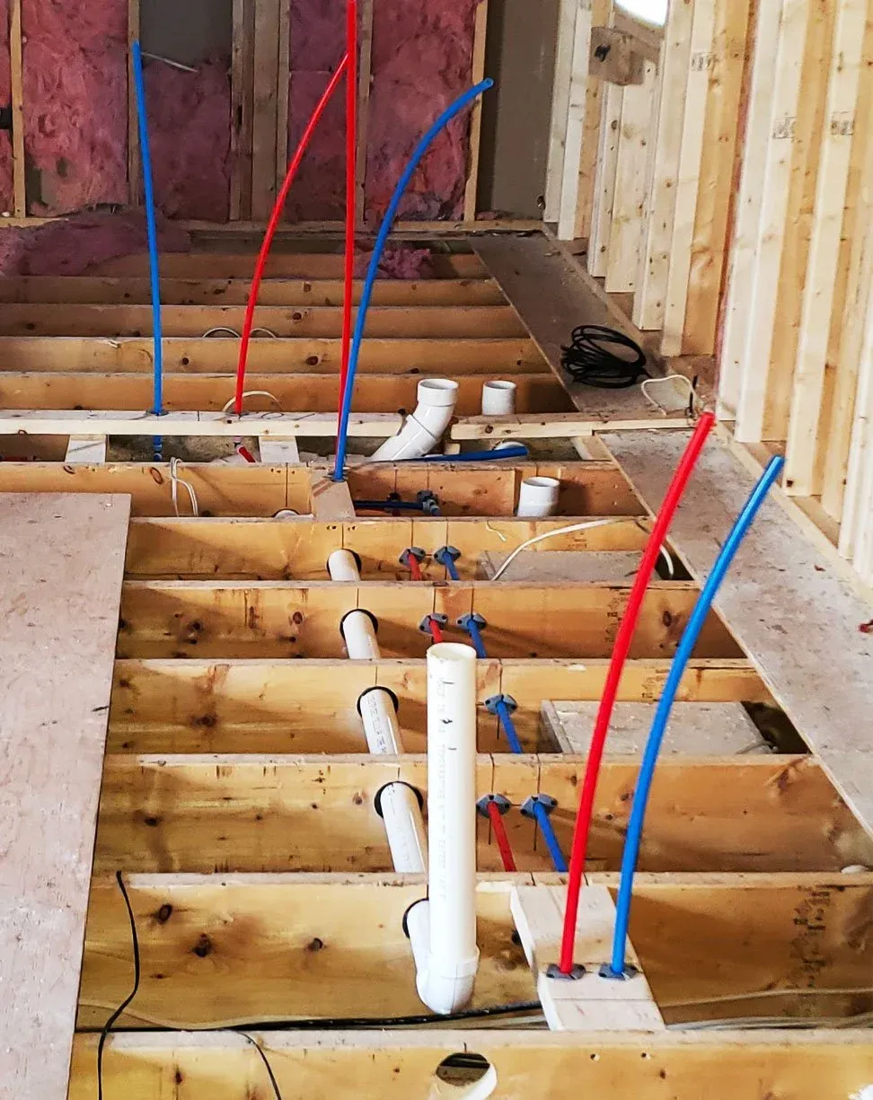 Construction site showing wooden floor framing with various red, blue, and white pipes and tubing for plumbing, electrical wiring, and HVAC systems.