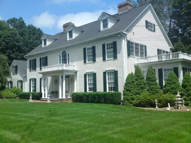Large white house with black shutters, multiple dormer windows, green manicured lawn, and landscaped garden with shrubs and small statues.
