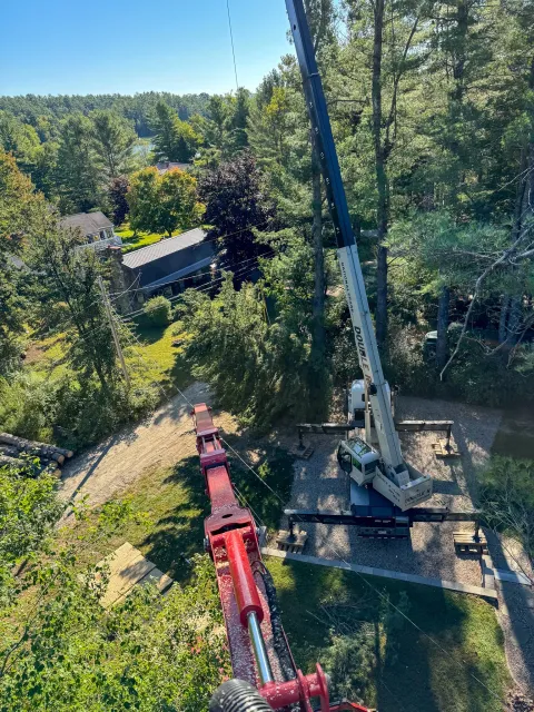 Aerial view of a tree trimming or maintenance operation using a large crane with an extended boom, positioned on a gravel area near trees and residential houses in a green, wooded neighborhood. Tree Service Maine, Learned Tree Service