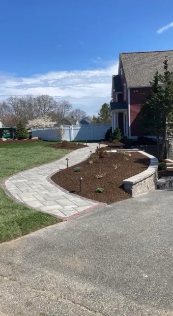 Paver walkway curves towards a red house with a brown mulch bed and retaining wall, blue sky.