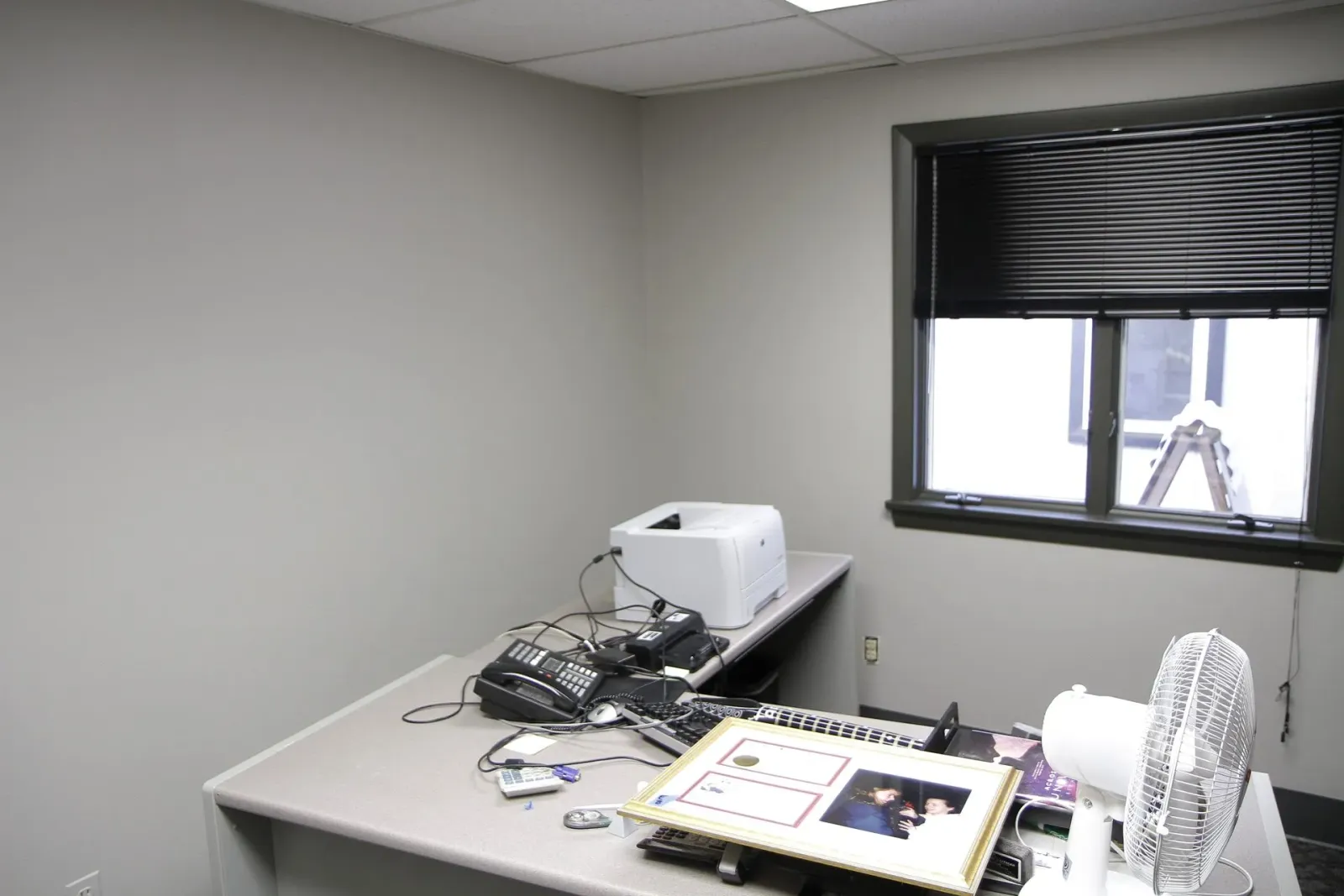 A desk with a printer and a fan in a room with a window.