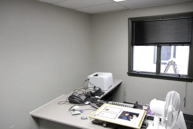 A desk with a printer and a fan in a room with a window.