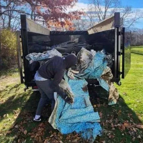 Person loading debris into a black dumpster outdoors. Blue tarp and fall foliage present.